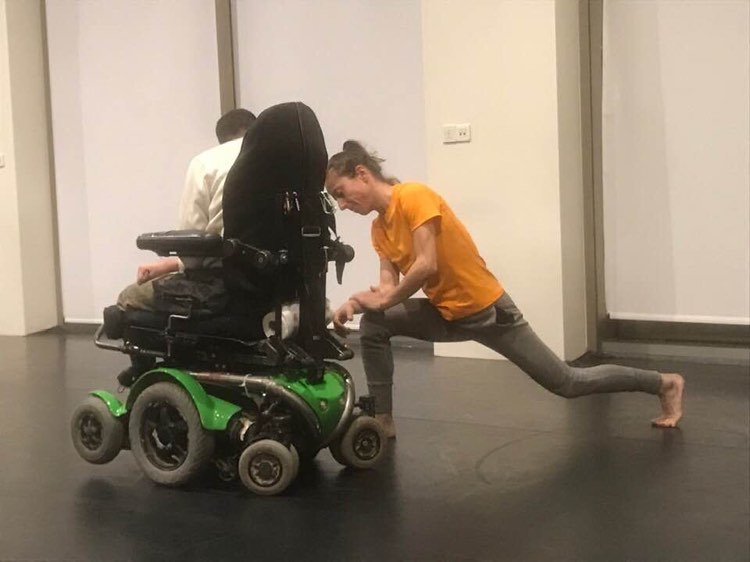 Maria, a female dancer, wearing an orange t-shirt leans towards Marios, a crip performer on his wheels