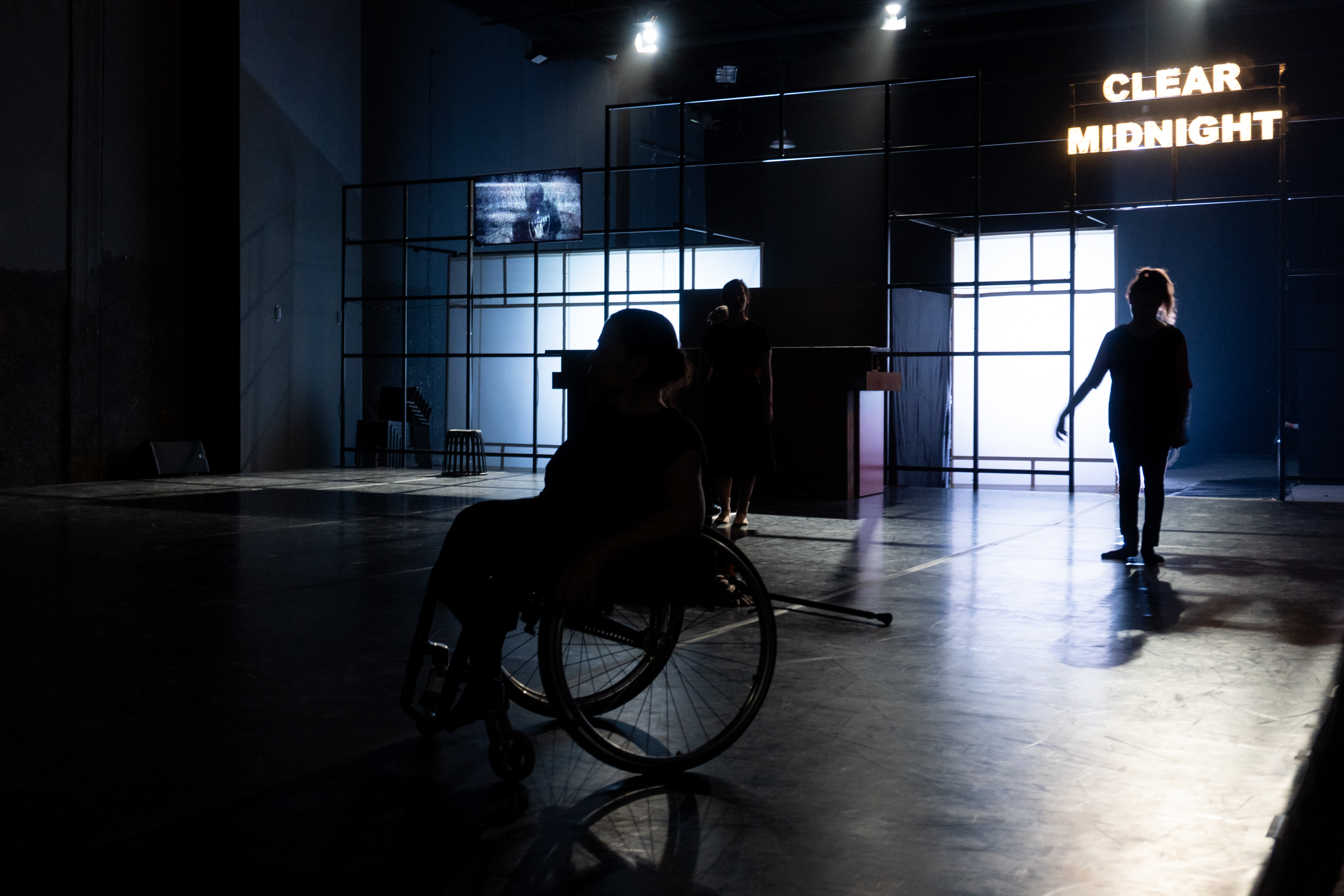 Three dancers, one in a wheelchair, are in front of a white set in a black box theatre setting