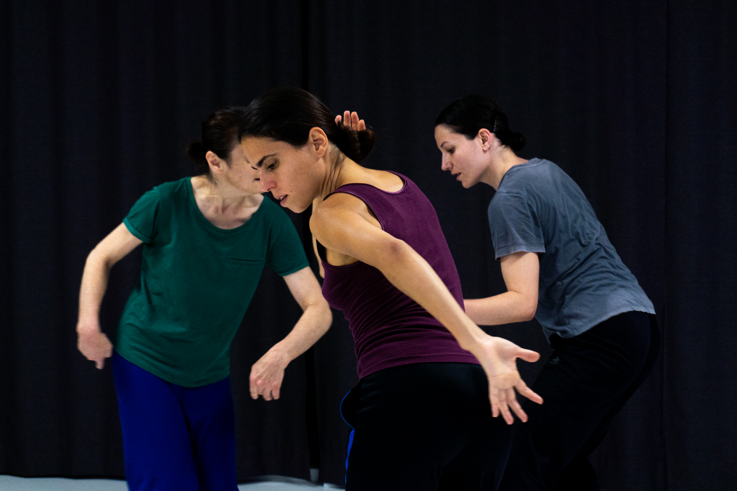 Three female performers in green, purple and grey t-shirts dance in proximity with their arms in different shapesr arms