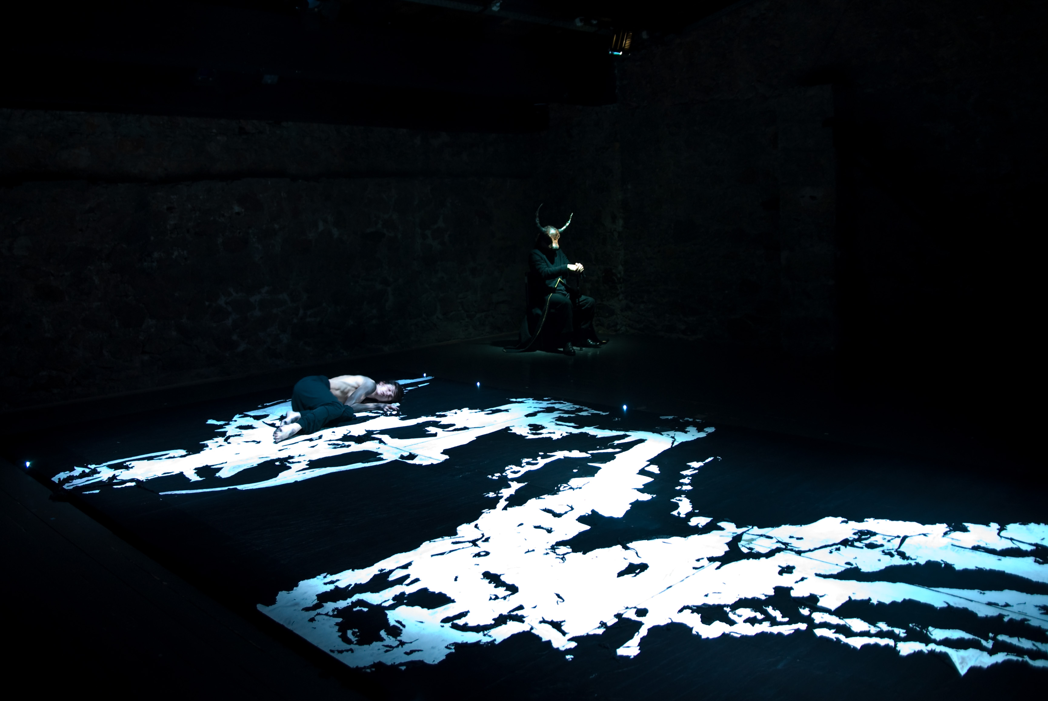 a white marked geography is formed on a black canvas floor with two performers one on the floor and the second on a chair with a mask of a minotaurus