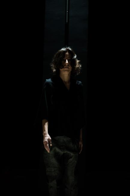 Maria, a performer dressed in black, looks front with her face and right arm prevailing