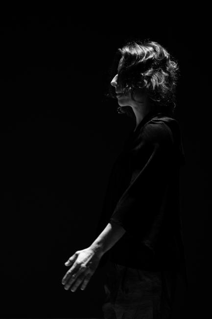 Maria, a performer dressed in black, faces left with her face and left arm prevailing