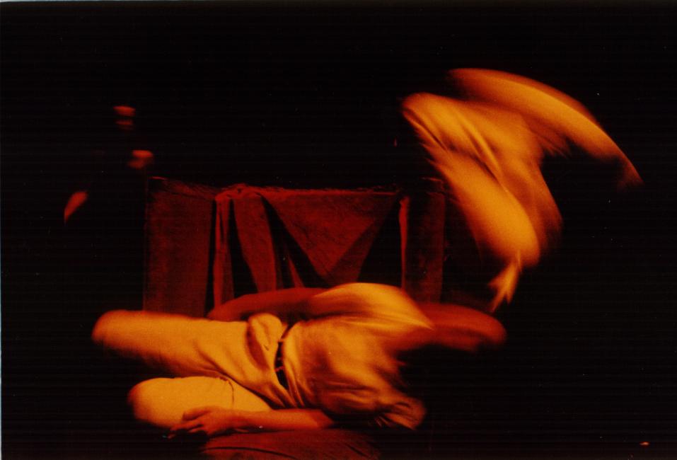 two performers on a red chair, the one horizontally and the other seen vaguely